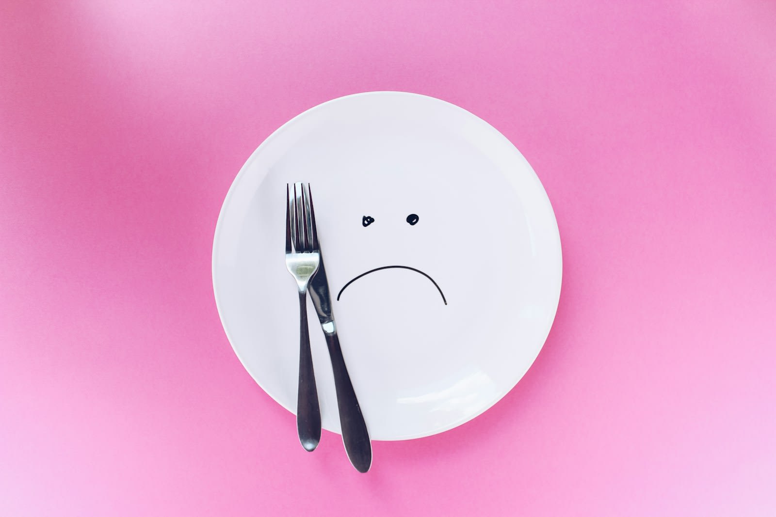 An empty white plate with a fork and knife on a pink background